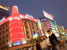 Busy Shanghai street with advertisements along the walls of the buildings. The most prominenet advert is a large lit-up Coca-cola bottle on the corner of a building.  (http://www.bbc.co.uk/staticarchive/be556f1cd99b5117fc23969de8923b7b1886560c.jpg)